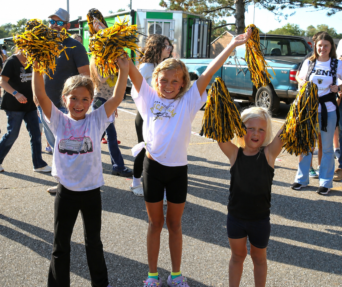 Three young girls in cheerleading uniforms holding pom poms, standing on a street next to a car and a truck.