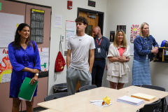 People standing in a classroom, posing for a picture, with some holding papers and a book.