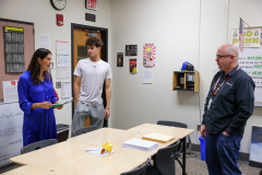 Two men and a woman in a classroom setting, standing near a table with papers on it, discussing something.