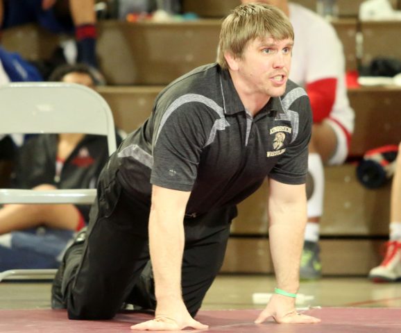 Man in black uniform kneeling on floor, leaning forward, hands resting on knees, looking down, intense focus.