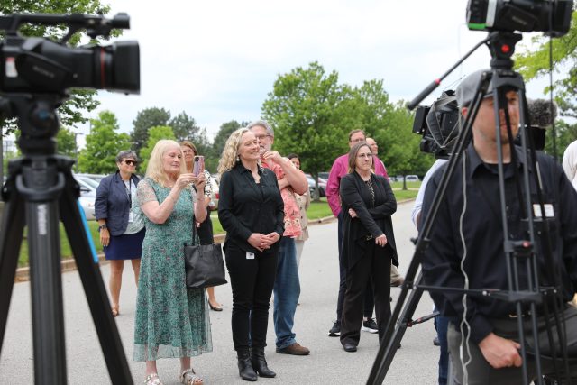 A group of people gathered around a news event, with a camera person filming and a cameraman operating a camera on a tripod.
