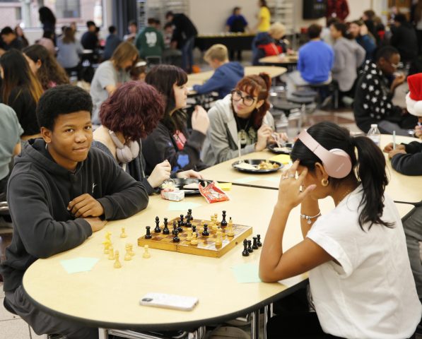 Group of people gathered around a dining table in a cafeteria setting, engaged in conversation and playing chess.