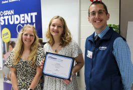 Student standing in middle holding C-SPAN award with teacher and man from C-SPAN alongside her