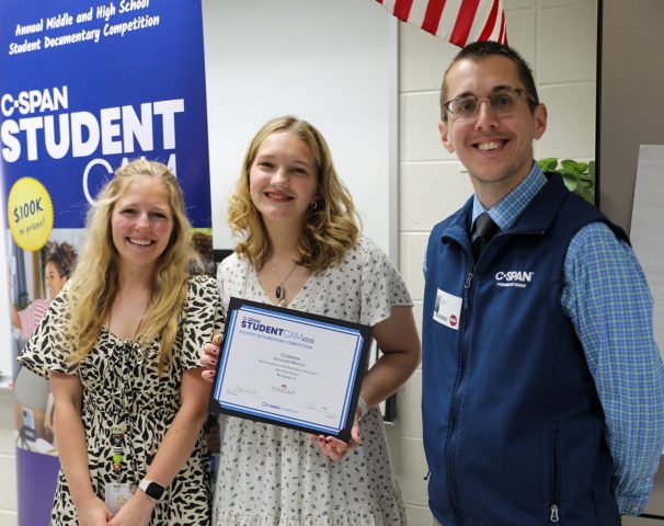 Student standing in middle holding C-SPAN award with teacher and man from C-SPAN alongside her