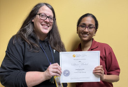 Caelea Armstrong and Sreeja Bhattacharjee pose in front of a yellow wall holding an award certificate between them.