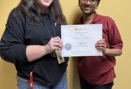 Caelea Armstrong and Sreeja Bhattacharjee pose in front of a yellow wall holding an award certificate between them.