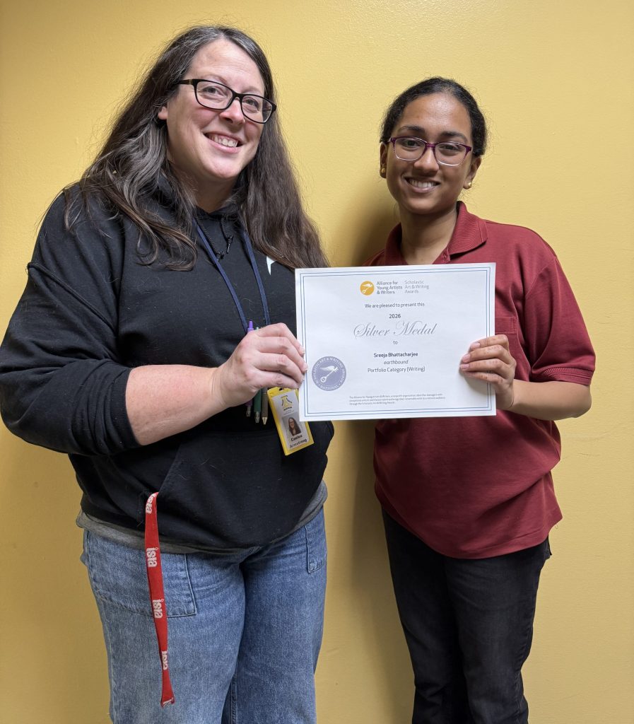 Caelea Armstrong and Sreeja Bhattacharjee pose in front of a yellow wall holding an award certificate between them.