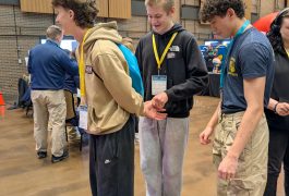 Three young men in a room, one holding hands with another while the third looks on, all wearing backpacks and casual