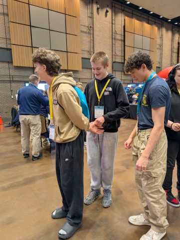 Three young men in a room, one holding hands with another while the third looks on, all wearing backpacks and casual