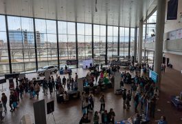 Large room filled with people standing around tables, some of which have laptops on them; a stage with a speaker at one end