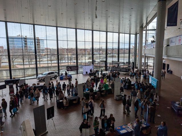 Large room filled with people standing around tables, some of which have laptops on them; a stage with a speaker at one end