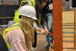 Two women in hard hats working on a construction site, one holding a hammer, standing next to a wooden structure.