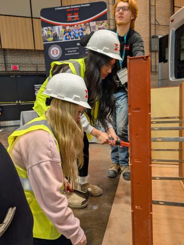 Two women in hard hats working on a construction site, one holding a hammer, standing next to a wooden structure.
