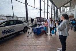 Woman standing in front of a car display, holding a microphone, addressing an audience.