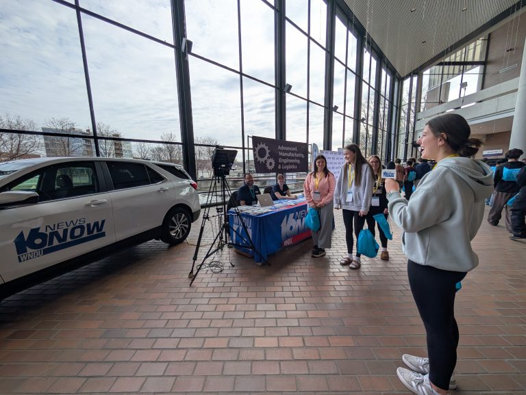 Woman standing in front of a car display, holding a microphone, addressing an audience.