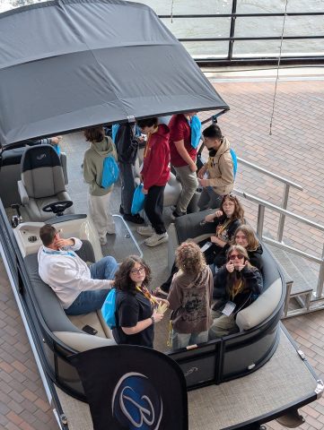 People inside a boat on display at a convention, gathered around an open roof area.