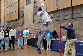 People in a gymnasium playing basketball. One person is jumping to catch the ball while others watch from the sidelines.