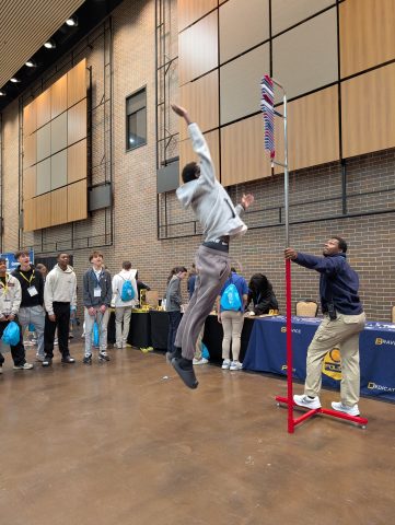 People in a gymnasium playing basketball. One person is jumping to catch the ball while others watch from the sidelines.