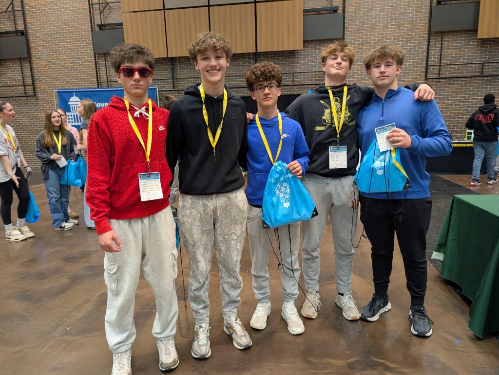Five young men posing together in a room, all wearing yellow lanyards around their necks and holding blue bags, standing in