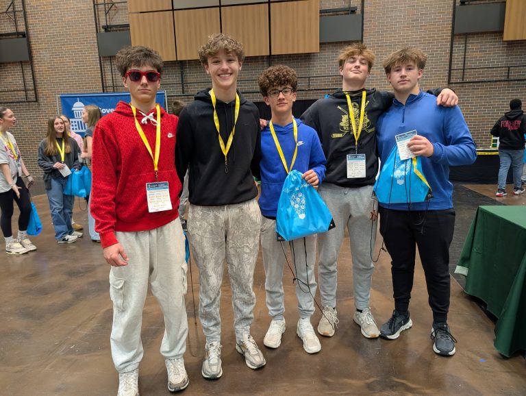 Five young men posing together in a room, all wearing yellow lanyards around their necks and holding blue bags, standing in
