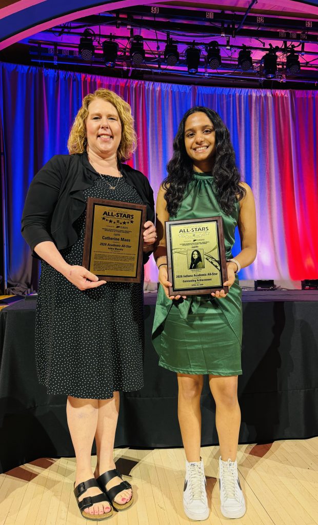 Two people standing on a stage holding awards. The person on the left is wearing a black dress and glasses, while the person