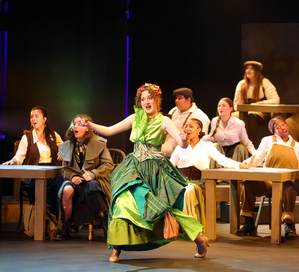 Theatrical performance of a woman in a green dress dancing on stage, surrounded by other performers and set against a