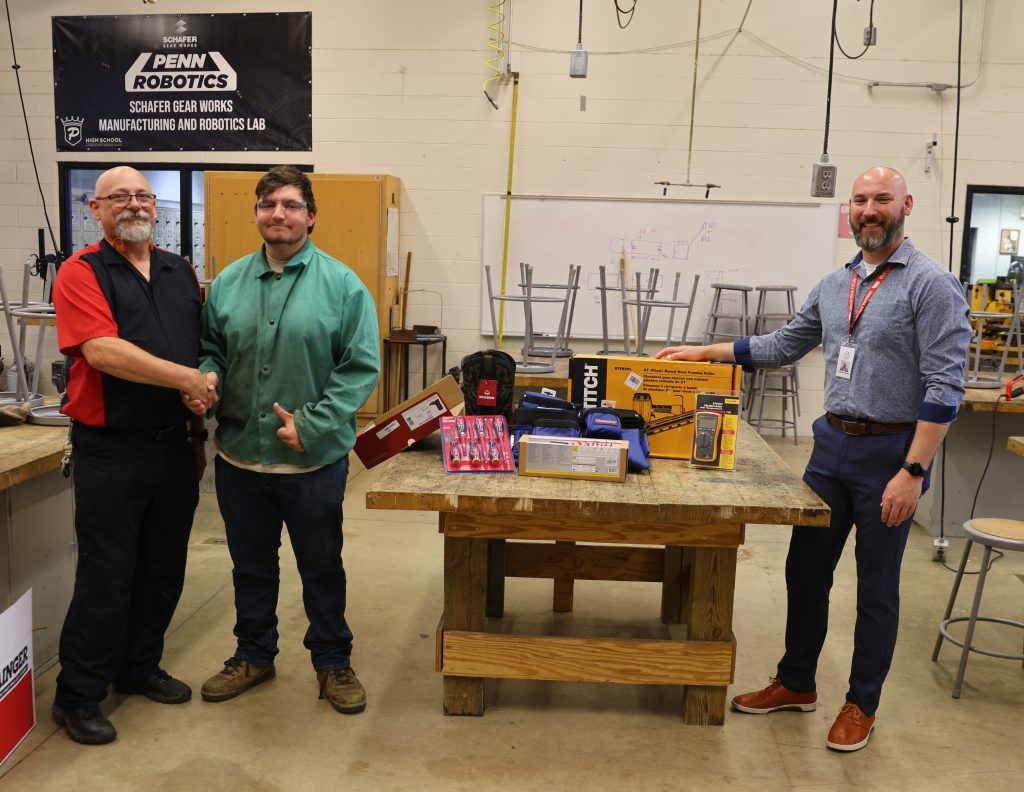 Three men standing in front of a table with various items on it, posing for a picture in a workshop setting.