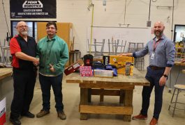 Three men standing in front of a table with various items on it, posing for a picture in a workshop setting.