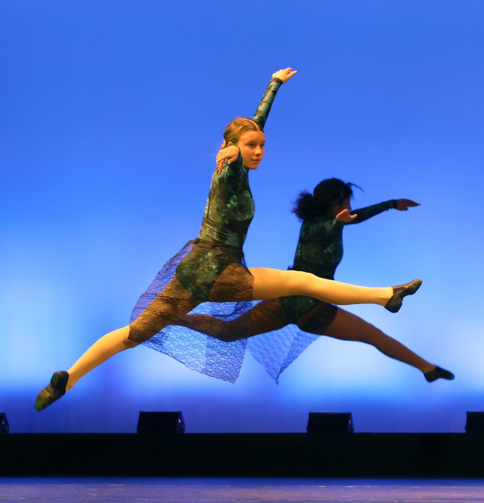 Two dancers performing ballet on stage, one in mid-air leap.