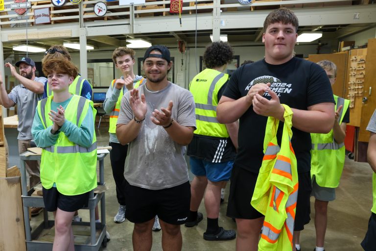 Group of people in a workshop setting, some clapping, others standing around, all wearing safety vests and hard hats, posing