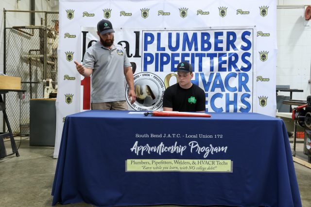 Two men at a table with a sign that reads "Plumbers Pipefitters WAC".