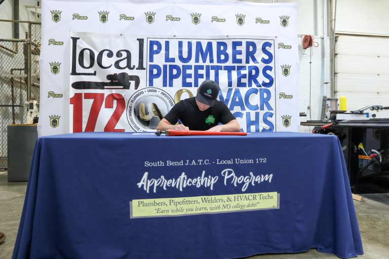 A person sitting at a table with a sign that reads 'Local Plumbers Pipefitters' and a banner in the background promoting an