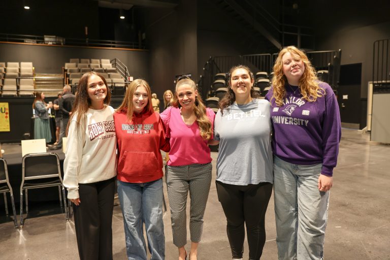 Students seated at a table in front of a podium, with a banner displaying the name of an educational institution behind them.