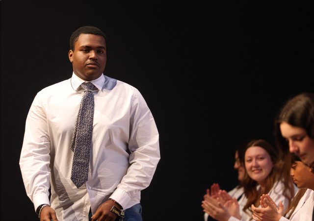 Man in a tie standing on stage, receiving applause from audience members.