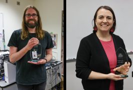Two people holding awards in a room, one person on left is bearded, both smiling and looking at the camera.