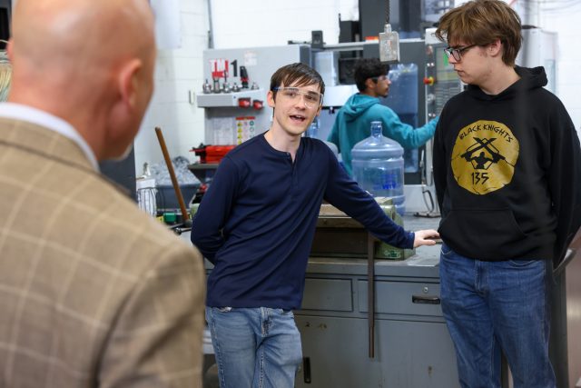 Two young men in a workshop setting, one leaning against a workbench with a drill press in the background, and another