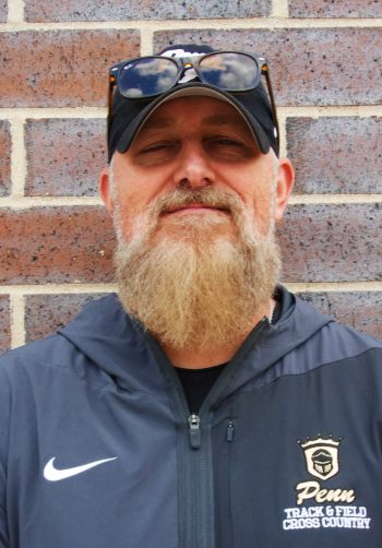 Man in beard posing for picture, smiling slightly, wearing baseball cap, glasses, jacket with logo and name of school,