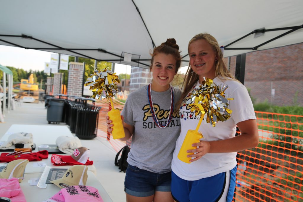 Two individuals posing in front of a tent at an outdoor event. The person on the left is holding a yellow pom-pom and