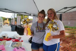 Two individuals posing in front of a tent at an outdoor event. The person on the left is holding a yellow pom-pom and