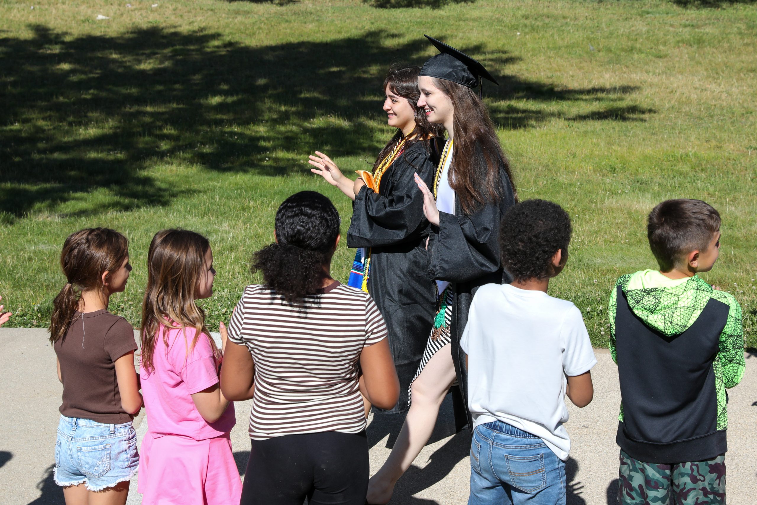 Students in graduation cap and gown walking near and waving younger students