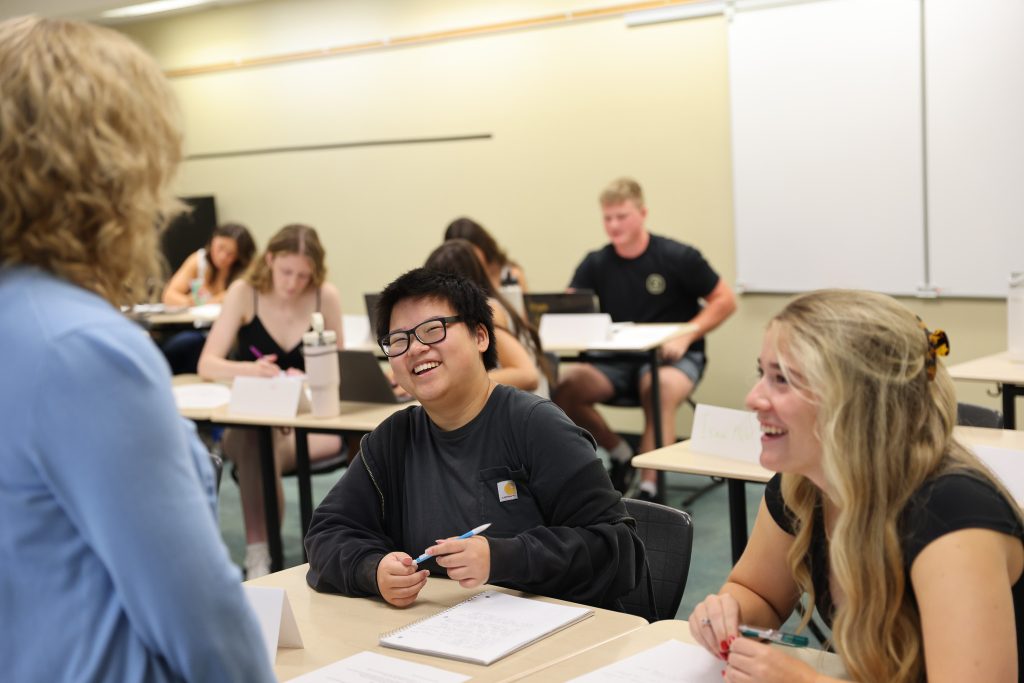 A woman stands in front of a group of students, smiling and addressing them, as they attentively listen and take notes.
