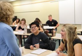 A woman stands in front of a group of students, smiling and addressing them, as they attentively listen and take notes.