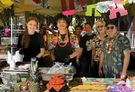 People gathered around a food stand with a tropical theme, including a pineapple and a hula girl sign.