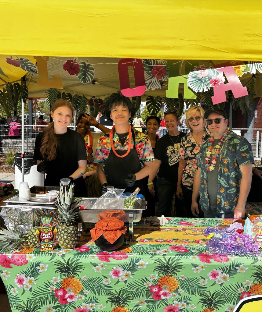 People gathered around a food stand with a tropical theme, including a pineapple and a hula girl sign.