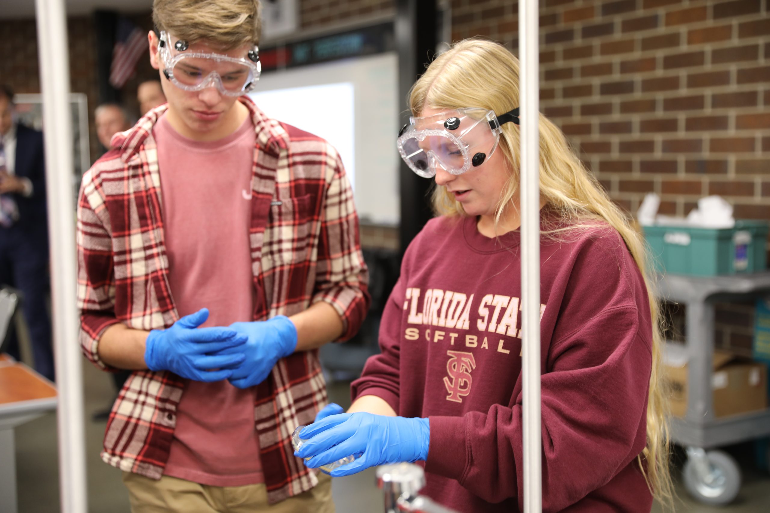 Two people in a lab, one wearing a red shirt and the other in a blue shirt, both wearing safety goggles and gloves, standing
