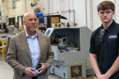 Two men in a factory setting, one standing and one sitting, engaged in conversation.