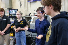 Group of people in a workshop, observing an object on a table. One person gestures while explaining something to others.