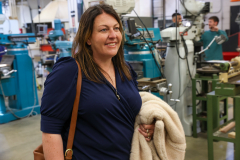 Woman standing in a workshop, smiling at camera, wearing blue shirt and brown purse.