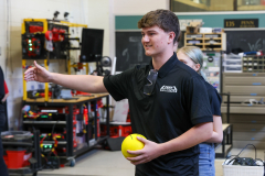 Man in black shirt holding a yellow ball in his hand, standing in front of shelves filled with various items and machinery.
