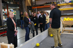 People gathered in a workshop, observing a man preparing to play darts.
